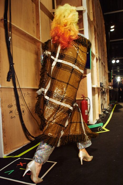 Editorial fashion image of model in patterned coat with red and yellow hair, backstage.