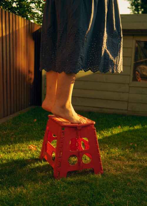 Lifestyle image of bare feet standing on a red stool in a garden.