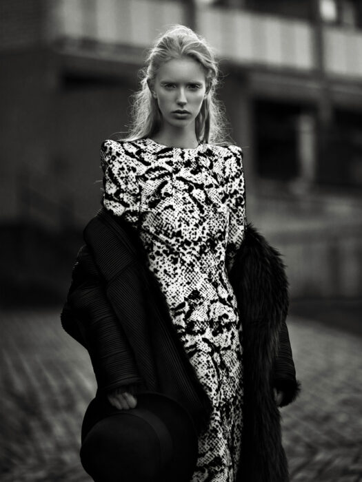 Black-and-white fashion editorial image of woman in printed dress and fur coat on a street.