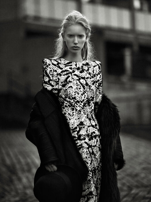 Black-and-white fashion editorial image of woman in printed dress and fur coat on a street.