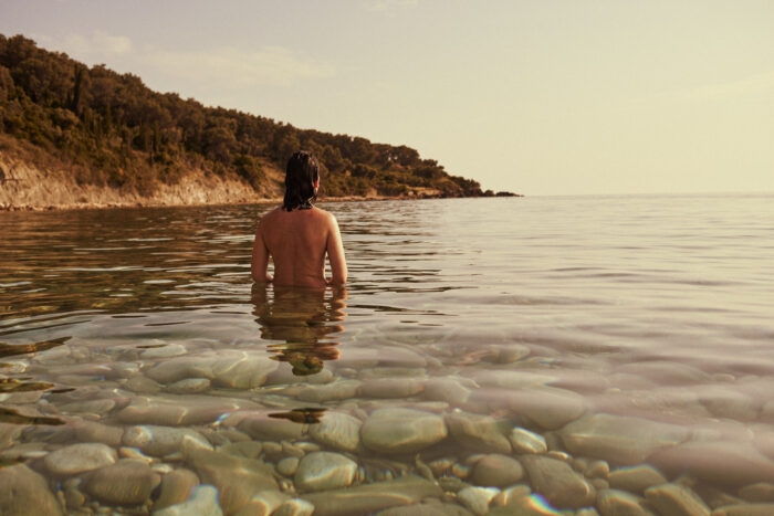 Travel portrait of a person standing in shallow sea along a rocky coastline.