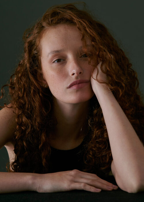 Portrait of woman with curly red hair leaning on her arm.